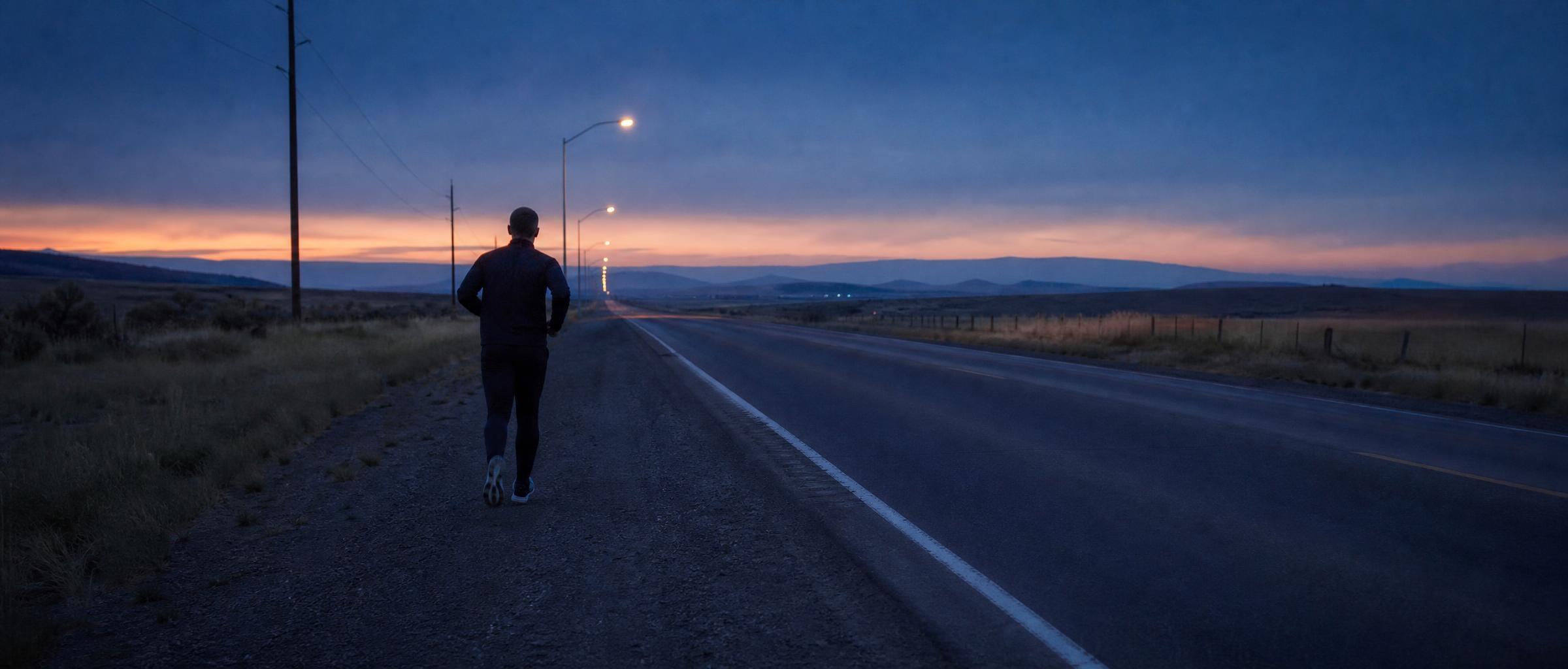 A man in his mid-forties, small in the frame, jogs the gravel shoulder beside an empty road running to the horizon at blue hour.