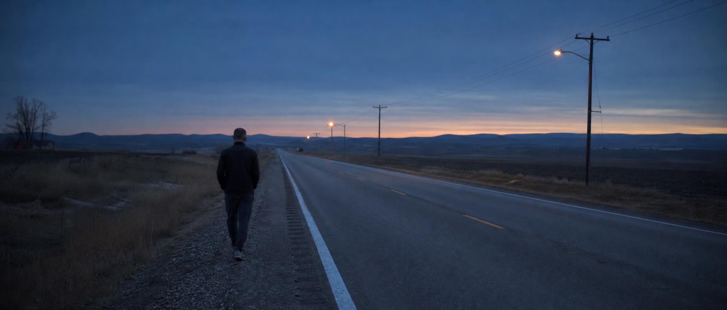 A man in his mid-forties, small in the frame, walks the gravel shoulder beside an empty road running to the horizon at blue hour.