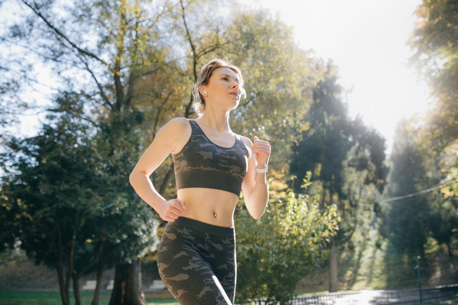 A woman runs through a sunlit park in autumn, a white watch on her wrist and an earbud in her ear, seen from slightly below.