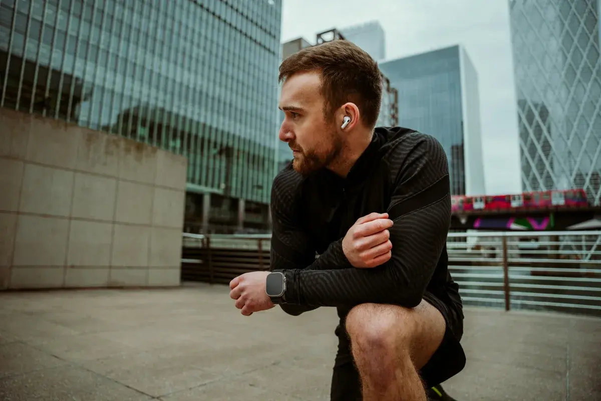 A man in black running gear crouched on a city plaza, a wireless earbud in his ear and a watch on his wrist.