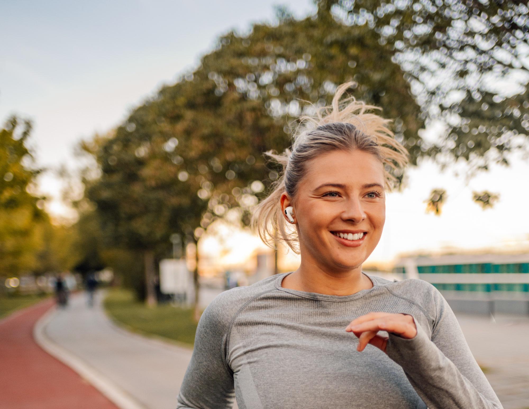 A smiling woman with a white earbud jogging toward the camera on a tree-lined park path in warm evening light.